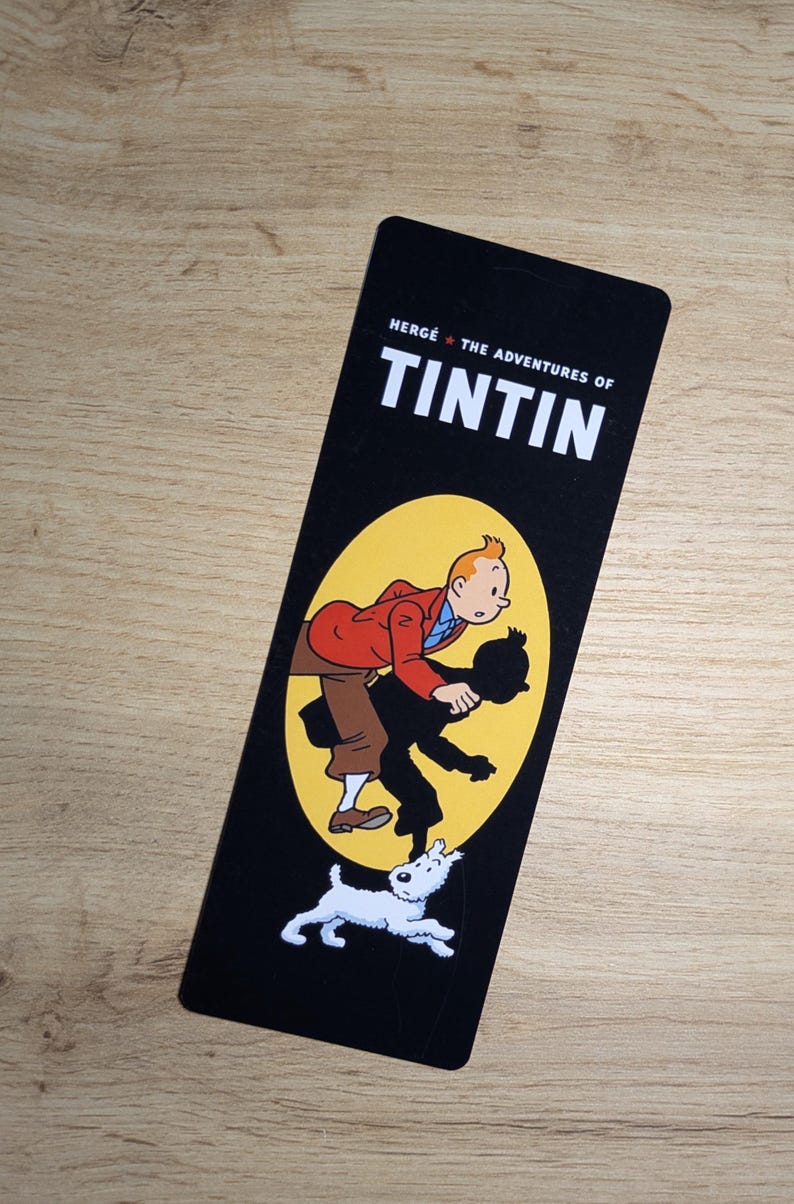May include: A rectangular black bookmark featuring the white text HERG&Eacute; THE ADVENTURES OF TINTIN at the top. Below the text, a yellow oval contains an illustration of Tintin in a red jacket and brown pants, with his black silhouette and Snowy the dog.
