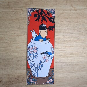 May include: A rectangular bookmark featuring a cartoon illustration of a person in a blue shirt and a small white dog emerging from a large white ceramic vase decorated with blue and pink floral patterns, a blue bird, and a vibrant red background.