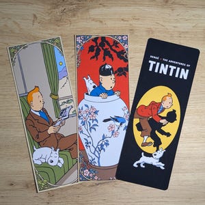 May include: Three rectangular paper bookmarks featuring Tintin and Snowy. The first shows Tintin reading on a green sofa. The second depicts Tintin inside a large blue and white floral vase. The third is black with the text The Adventures of Tintin.