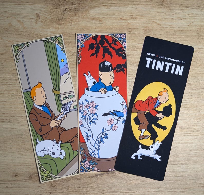 May include: Three rectangular paper bookmarks featuring Tintin and Snowy. The first shows Tintin reading on a green sofa. The second depicts Tintin inside a large blue and white floral vase. The third is black with the text The Adventures of Tintin.