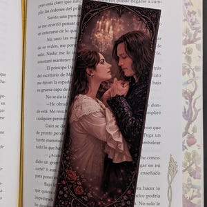 Puede incluir: Marcapáginas decorativo con una imagen romántica de una pareja, de estilo vintage. El marcapáginas tiene un borde oscuro con detalles florales y está colocado dentro de un libro con texto visible.