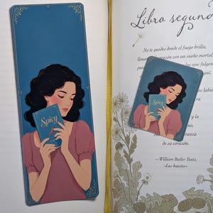 Puede incluir: Un marcapáginas rectangular y una tarjeta cuadrada con la ilustración de una persona de pelo oscuro con una blusa malva, sosteniendo un libro azul con la palabra Spicy y un icono de chile rojo en la portada, con detalles florales dorados.