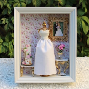 Puede incluir: Un vestido de novia blanco en un maniquí en una caja de sombra con un fondo floral. Un pequeño pastel de bodas con las palabras "we do" está en una mesa a la izquierda. Una foto enmarcada de una novia y un novio está en la pared a la derecha.