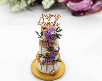 Puede incluir: Un pastel blanco de dos pisos con glaseado dorado y flores moradas y blancas. El pastel está decorado con macarons y tiene un adorno que dice "mr & mrs Dukecki".