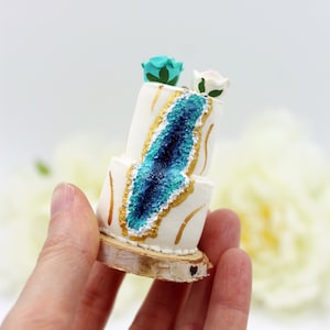 May include: A miniature wedding cake with a blue and gold geode design, topped with two small flowers. The cake is on a wooden base.