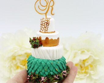 Puede incluir: Un pastel de boda de tres pisos con un diseño dorado y blanco. El nivel inferior está cubierto de fondant verde que se asemeja a escamas. El pastel está coronado con una letra "R" dorada y un pequeño ramo de flores rojas y verdes.