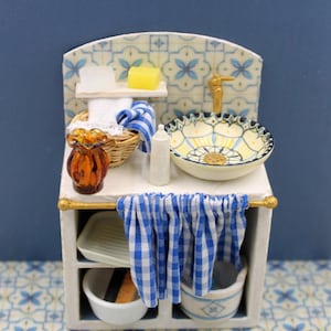 Puede incluir: Mini fregadero blanco con un salpicadero con estampado azul y blanco. El fregadero tiene un grifo dorado, un cuenco decorativo y una toalla a cuadros azules y blancos. También hay jabones, toallas y una cesta en miniatura.