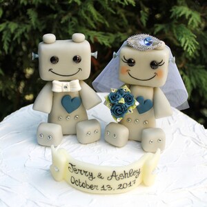 Pu&ograve; includere: Un topper per torta nuziale a forma di robot grigio con un cuore blu e un papillon bianco. La sposa robot indossa un velo bianco e una corona di gioielli blu. Il topper per torta ha uno striscione che recita "Terry & Ashley October 13, 2017".