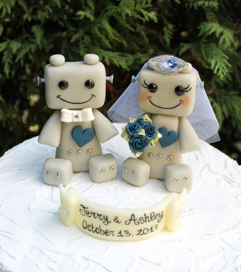 Pu&ograve; includere: Un topper per torta nuziale a forma di robot grigio con un cuore blu e un papillon bianco. La sposa robot indossa un velo bianco e una corona di gioielli blu. Il topper per torta ha uno striscione che recita "Terry & Ashley October 13, 2017".
