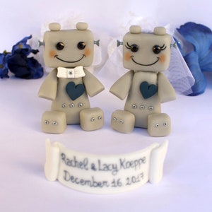 Pu&ograve; includere: Due figurine di robot grigi vestiti da sposa e sposo. La sposa ha un velo bianco e lo sposo un papillon bianco. Entrambi i robot hanno un cuore blu sul petto. Lo striscione sotto di loro recita "Rachel & Lacy Koepe December 16, 2017".