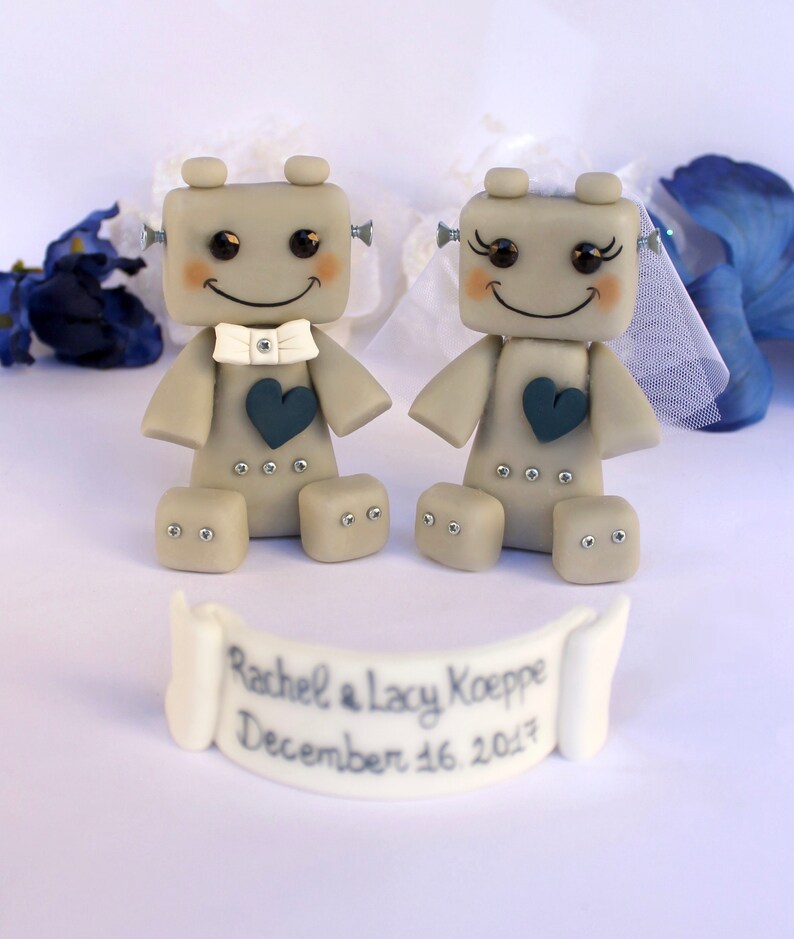 Pu&ograve; includere: Due figurine di robot grigi vestiti da sposa e sposo. La sposa ha un velo bianco e lo sposo un papillon bianco. Entrambi i robot hanno un cuore blu sul petto. Lo striscione sotto di loro recita "Rachel & Lacy Koepe December 16, 2017".