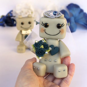 Pu&ograve; includere: Una coppia di figurine di robot vestite da sposa e sposo. La sposa indossa un velo bianco e una corona di gioielli blu. Tiene un bouquet di rose blu-verdi. Lo sposo indossa un papillon bianco.
