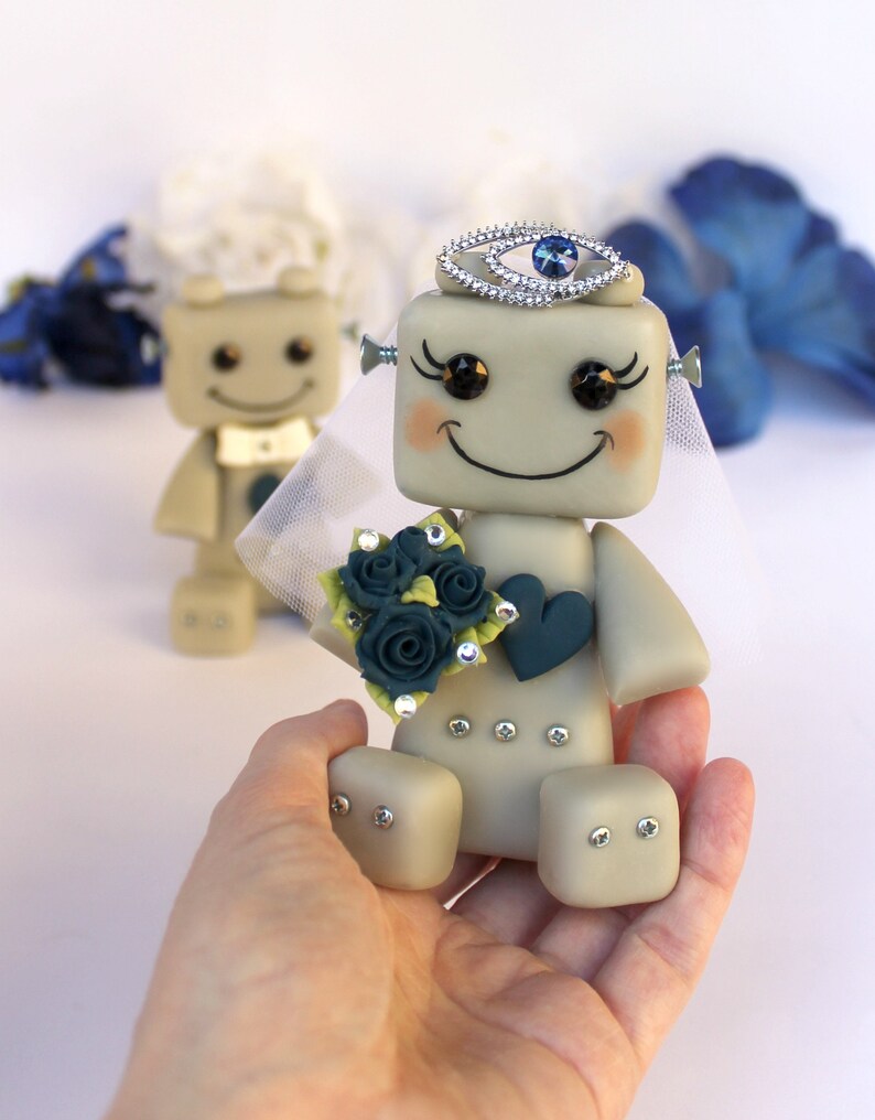 Pu&ograve; includere: Una coppia di figurine di robot vestite da sposa e sposo. La sposa indossa un velo bianco e una corona di gioielli blu. Tiene un bouquet di rose blu-verdi. Lo sposo indossa un papillon bianco.