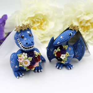 May include: Blue dinosaur and pterodactyl wedding cake toppers with gold crowns and floral bouquets. The dinosaur is wearing a white veil.