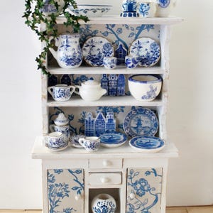 Könnte beinhalten: Ein weißer Miniaturschrank mit blau-weißen Dekorationen im Delfter Stil. Der Schrank ist mit winzigen Tellern, Tassen, Teekannen und einer Windmühlenfigur gefüllt. Eine kleine grüne Pflanze sitzt obenauf und verleiht dem Display eine natürliche Note.