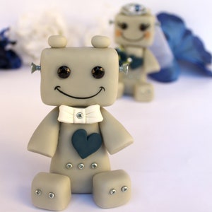Pu&ograve; includere: Una statuetta di robot grigio con un papillon bianco e un cuore blu, sorridente con occhi neri. Il robot &egrave; seduto su una superficie bianca.
