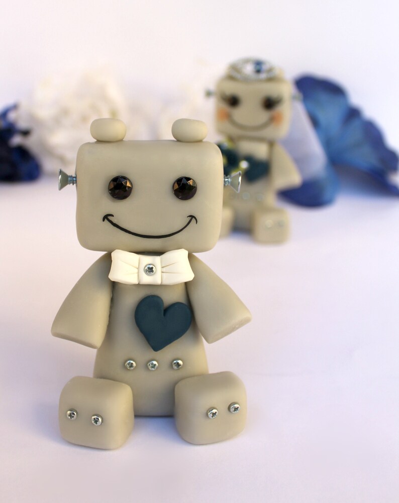 Pu&ograve; includere: Una statuetta di robot grigio con un papillon bianco e un cuore blu, sorridente con occhi neri. Il robot &egrave; seduto su una superficie bianca.