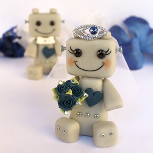 Pu&ograve; includere: Una coppia di figurine da matrimonio di robot. La sposa robot indossa un velo bianco e una corona con un gioiello blu. Tiene in mano un bouquet di fiori blu e verdi. Lo sposo robot indossa un papillon bianco.