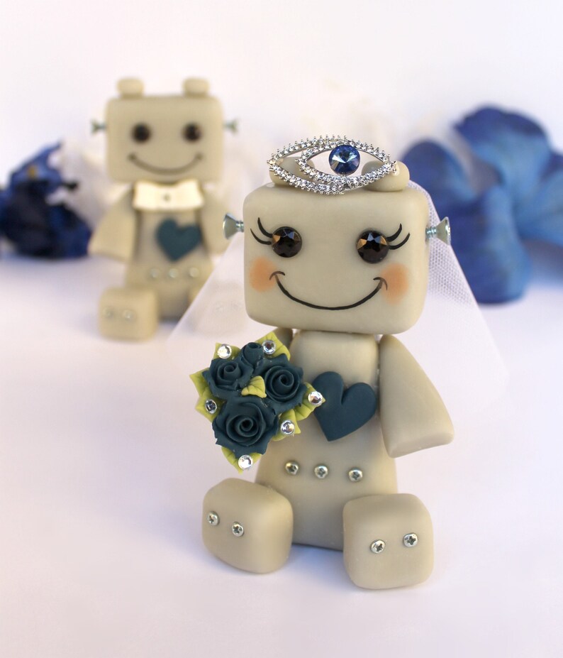 Pu&ograve; includere: Una coppia di figurine da matrimonio di robot. La sposa robot indossa un velo bianco e una corona con un gioiello blu. Tiene in mano un bouquet di fiori blu e verdi. Lo sposo robot indossa un papillon bianco.