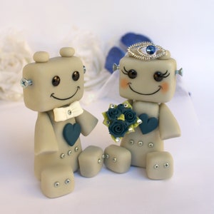 Pu&ograve; includere: Un topper per torta nuziale a forma di robot grigio con un papillon bianco e un cuore blu. La sposa robot indossa un velo bianco e una corona blu con un gioiello blu. Tiene un bouquet di fiori blu e verdi.