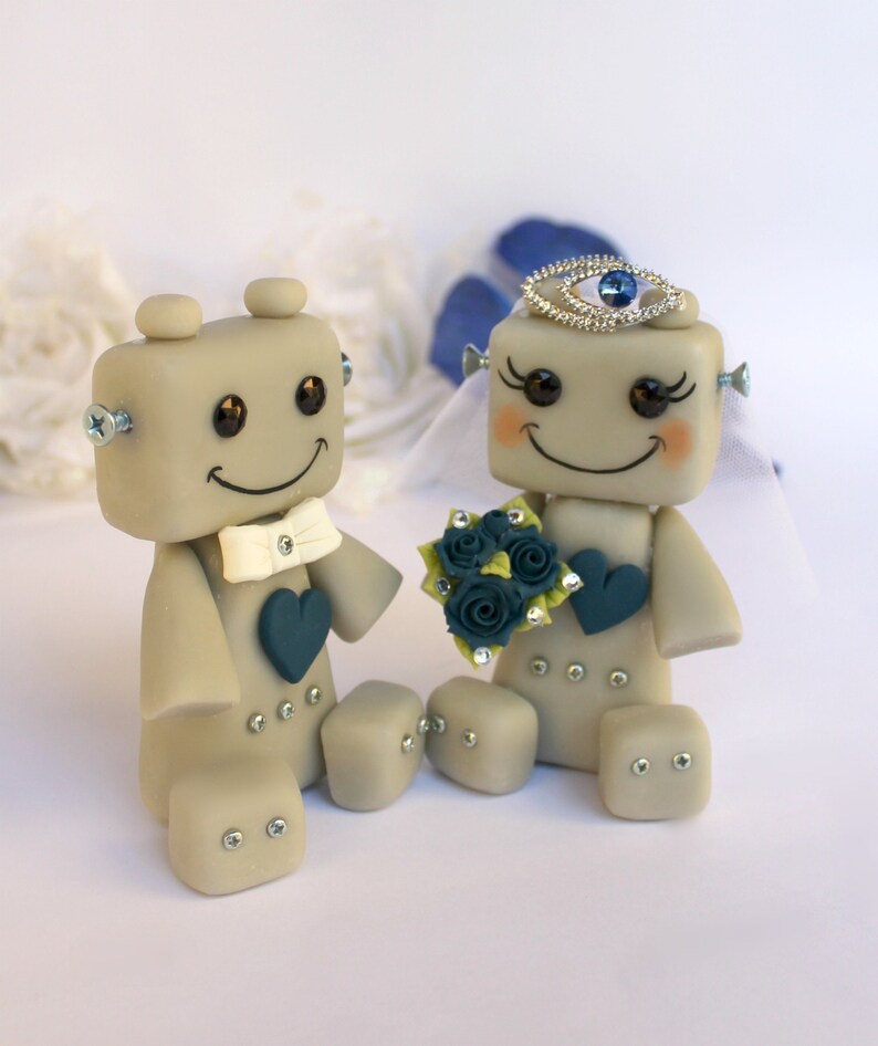 Pu&ograve; includere: Un topper per torta nuziale a forma di robot grigio con un papillon bianco e un cuore blu. La sposa robot indossa un velo bianco e una corona blu con un gioiello blu. Tiene un bouquet di fiori blu e verdi.