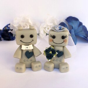 Pu&ograve; includere: Una coppia di topper per torta nuziale a forma di robot grigi. La sposa robot indossa un velo bianco e una tiara argentata con un gioiello blu. Lo sposo robot indossa un papillon bianco. Entrambi sorridono e tengono un bouquet di fiori blu scuro.