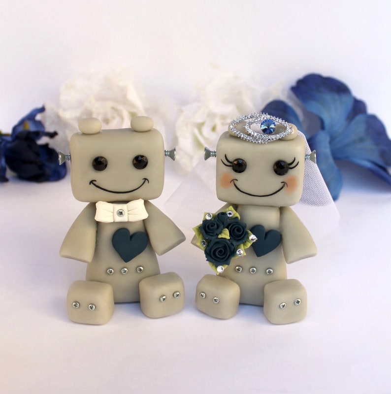Pu&ograve; includere: Una coppia di topper per torta nuziale a forma di robot grigi. La sposa robot indossa un velo bianco e una tiara argentata con un gioiello blu. Lo sposo robot indossa un papillon bianco. Entrambi sorridono e tengono un bouquet di fiori blu scuro.