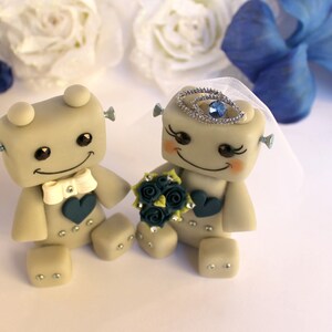 Pu&ograve; includere: Una coppia di figurine di robot grigi vestiti da sposa e sposo. La sposa indossa un velo bianco e una corona di gioielli blu. Lo sposo indossa un papillon bianco. La sposa tiene un bouquet di fiori blu e verdi.