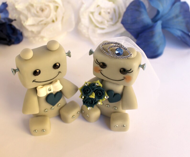 Pu&ograve; includere: Una coppia di figurine di robot grigi vestiti da sposa e sposo. La sposa indossa un velo bianco e una corona di gioielli blu. Lo sposo indossa un papillon bianco. La sposa tiene un bouquet di fiori blu e verdi.