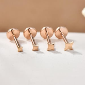 May include: Four rose gold stud earrings with a flat, round top and a star-shaped or square-shaped post. The earrings are displayed on a white surface with a soft, neutral background. The earrings are small and delicate.