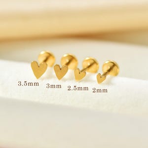 Könnte beinhalten: Vier goldfarbene herzförmige Ohrstecker mit flachem Verschluss, die in einer Reihe von der größten zur kleinsten angeordnet sind. Die Größen sind mit 3,5 mm, 3 mm, 2,5 mm und 2 mm beschriftet und geben die Breite jedes Herzmotivs an.