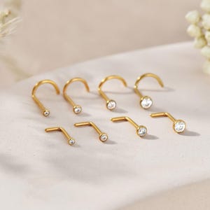 May include: A selection of gold-coloured nose rings. Some are curved, and others are straight, each with a small, clear gemstone. The jewellery is displayed on a light surface, with a soft, neutral background.