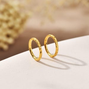 Könnte beinhalten: Ein Paar goldfarbene Creolen. Jeder Ohrring hat ein strukturiertes, geometrisches Muster. Die Ohrringe werden auf einer weißen Oberfläche mit einem weichen, verschwommenen Hintergrund präsentiert. Die Ohrringe sind rund und scheinen aus Metall zu sein.