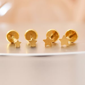 Könnte beinhalten: Vier goldfarbene Stern-Ohrstecker. Jeder Ohrring hat einen kleinen Sternanhänger, der an einer flachen Scheibe befestigt ist. Die Ohrringe werden auf einer hellen Oberfläche mit einem weichen, verschwommenen Hintergrund präsentiert.