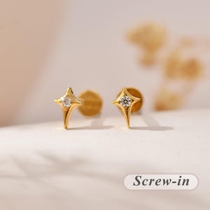 Könnte beinhalten: Ein Paar sternförmige Ohrstecker in Goldoptik, jeweils mit einem zentralen klaren Edelstein und einem Schraubverschluss. Der Text "Screw-in" ist in der unteren rechten Ecke des Bildes zu sehen und gibt den Verschlussstil des Schmucks an.