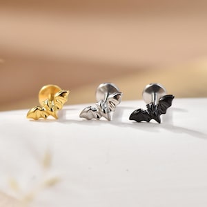 Könnte beinhalten: Drei Ohrstecker in Fledermausform, jeweils mit einem flachen Verschluss. Das Set enthält eine goldfarbene, eine silberfarbene und eine schwarze Fledermaus-Ohrstecker aus Metall, die alle in einer Reihe auf einer glatten, hellen Oberfläche liegen.