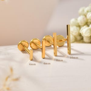 May include: Four gold-toned metal stud earrings with flat circular backings and vertical bar fronts. The earrings are arranged in a row, labelled with text indicating lengths of 4mm, 6mm, 8mm, and 10mm, showing the varying sizes of the posts.
