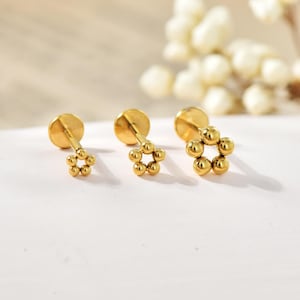 May include: Three gold stud earrings with a flower design. Each earring has a small, round disc and a cluster of small, spherical beads arranged in a floral pattern. The earrings are displayed on a white surface.