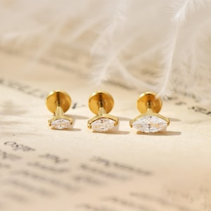 Könnte beinhalten: Drei goldfarbene Ohrstecker mit klaren, marquiseförmigen Zirkonia-Steinen. Die Ohrringe werden auf einer hellen Oberfläche mit einem weichen, verschwommenen Hintergrund und einer Feder präsentiert.