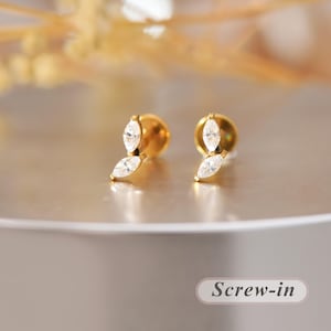 May include: A pair of gold-tone screw-in stud earrings, each featuring two marquise-cut clear stones arranged in a vertical leaf-like pattern. The earrings have a polished metal finish and a secure backing mechanism visible in the design.