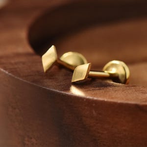 Peut inclure: Une paire de boucles d'oreilles dorées. Chaque boucle d'oreille présente une petite face en forme de losange et un dos rond. Les boucles d'oreilles sont présentées sur une surface en bois, mettant en valeur leur éclat métallique.