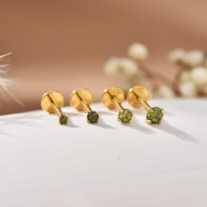 May include: Four gold-coloured stud earrings, each with a round, olive-green gemstone. The earrings are arranged in a row, with the gemstones increasing in size. They are set on a white surface with a blurred background.