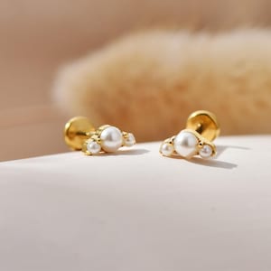 Könnte beinhalten: Zwei goldfarbene Ohrstecker, jeweils mit einer großen zentralen Perle und zwei kleineren Perlen. Die Ohrringe haben einen Stecker- und Verschluss. Der Hintergrund ist eine weiche, verschwommene Mischung aus Beige und Weiß.