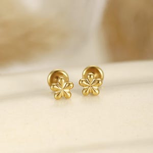 May include: A pair of gold-toned stud earrings shaped like five-petaled flowers. Each earring features a smooth, rounded finish with a central star-like indentation and a flat circular backing post, presenting a polished metallic surface throughout.
