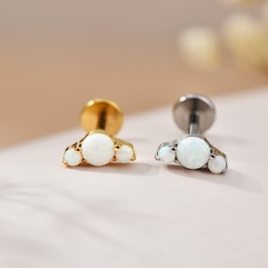 May include: Two pieces of jewellery, one gold and one silver, each with three white opal stones. The opals are arranged in a fan shape, with a larger central stone and two smaller ones. The jewellery is likely for ear piercings.