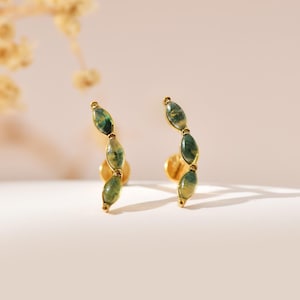 May include: A pair of gold-toned ear climbers featuring a curved design set with three marquise-shaped green moss agate stones. Each earring follows a vertical, slightly arched silhouette, showcasing the natural inclusions within the stones.