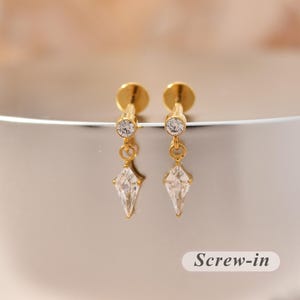 Könnte beinhalten: Ein Paar goldfarbene Schraubohrringe mit einem kleinen runden Stecker und einem klaren, facettierten, drachenförmigen Kristallanhänger, der an einem dünnen Goldring hängt. Der Text "Screw-in" ist in der unteren rechten Ecke zu sehen.