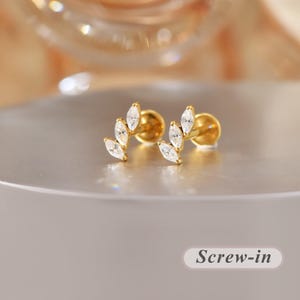 Könnte beinhalten: Ein Paar goldfarbene Ohrstecker mit geschwungenem Design, besetzt mit drei klaren, marquise-geschliffenen Steinen in Blattform. Die Ohrringe haben einen flachen, runden Verschluss und einen Schraubmechanismus. Der Text Screw-in ist sichtbar.