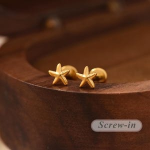 May include: A pair of gold-toned stud earrings shaped like five-pointed starfish. Each earring features a smooth, metallic finish and a screw-in backing mechanism. The text "Screw-in" is clearly visible in the lower right corner of the image.