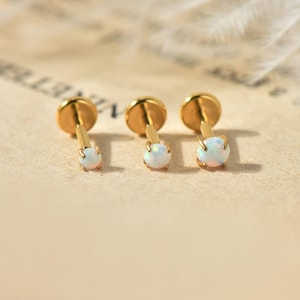 May include: Three gold-coloured stud earrings with opal-like, round gemstones. The earrings are displayed on a light brown surface. The earrings have a flat, circular backing and a post with a gemstone at the end.